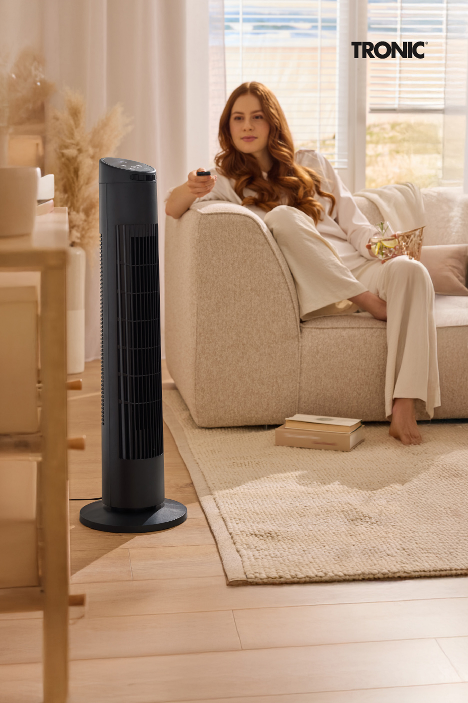 Een vrouw ontspant op de bank met een torenventilator ernaast, in een lichte woonkamer.