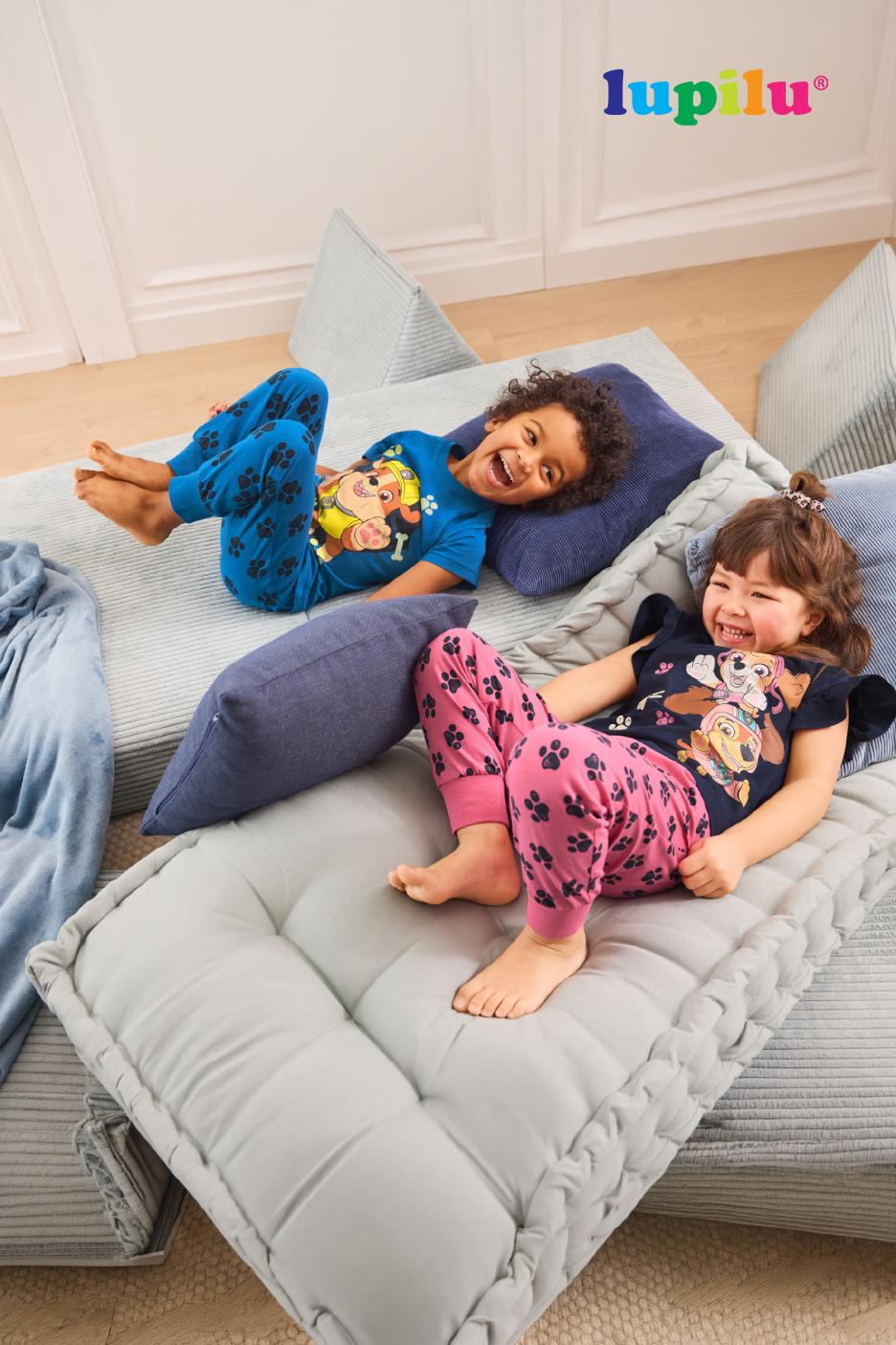 Twee lachende kinderen in Paw Patrol pyjama's liggen op grijze vloerkussens.