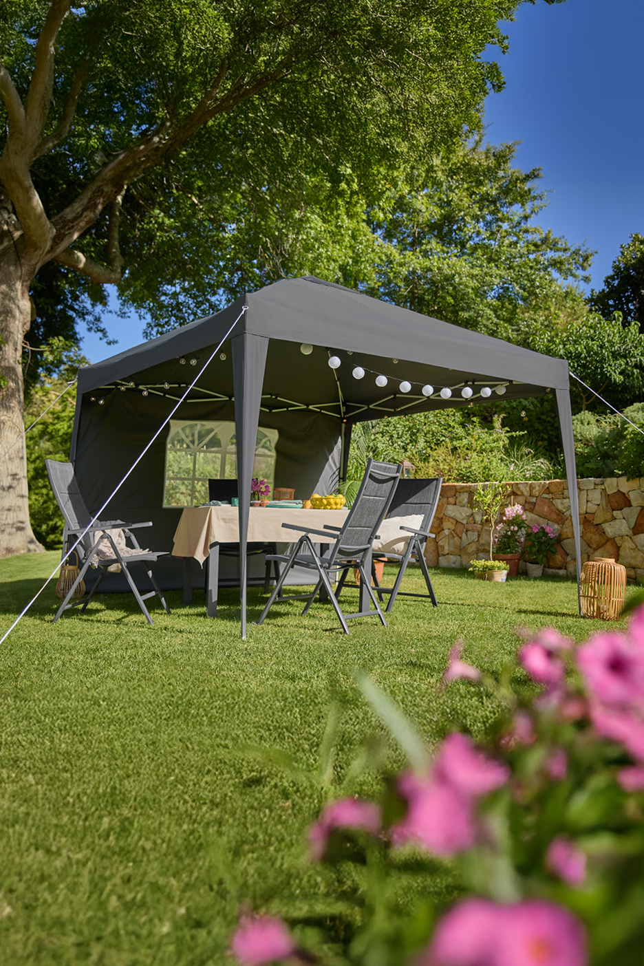 Grijze tuinpaviljoen met tafel en stoelen, versierd met lichtslingers, op een gazon.