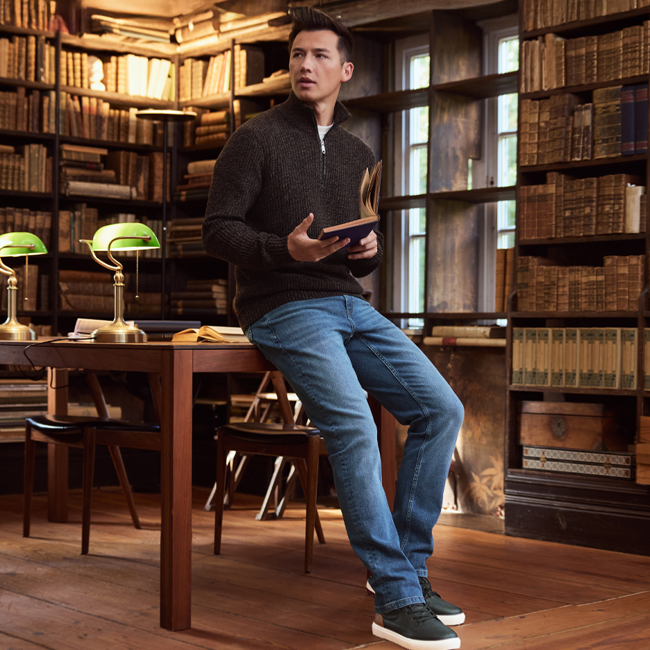 Man in donkerbruine trui en jeans leunt op een tafel in een bibliotheek.