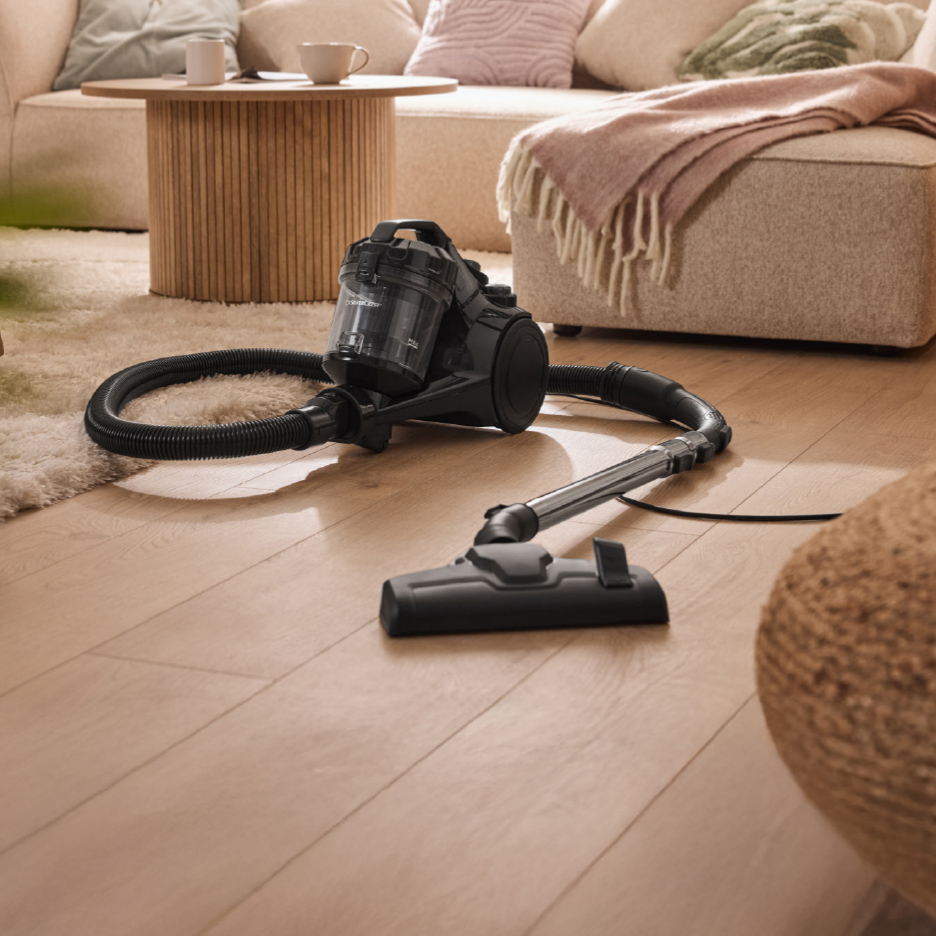 Zwarte stofzuiger op een houten vloer in een woonkamer met een beige bank.