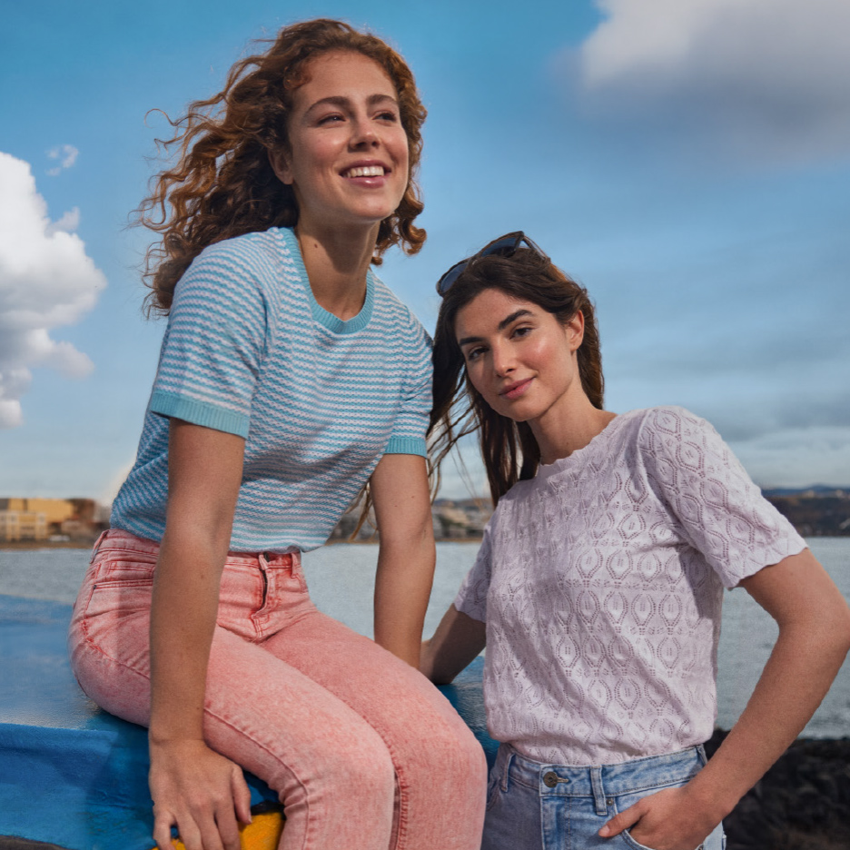 Twee lachende vrouwen in T-shirts en jeans, de een zittend, de ander staand, bij het water.