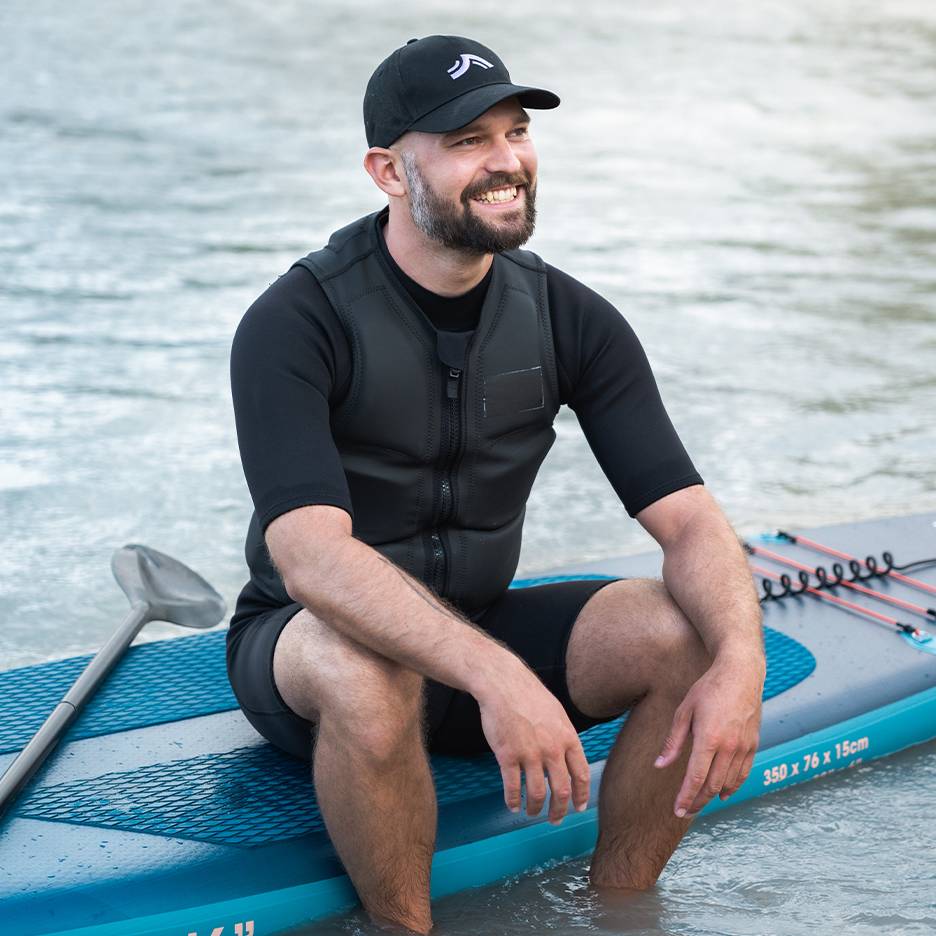 Lachende man met zwemvest en pet, zittend op een paddleboard in het water.