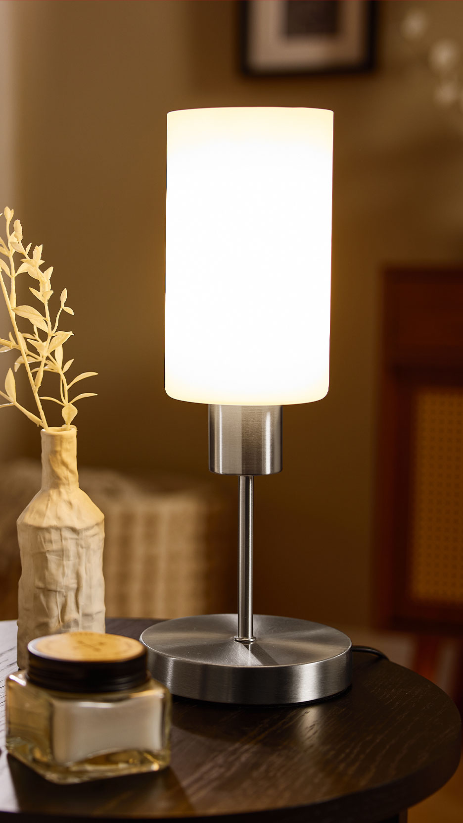 Moderne tafellamp met witte cilindrische kap en geborsteld metalen voet op een donkere houten tafel.