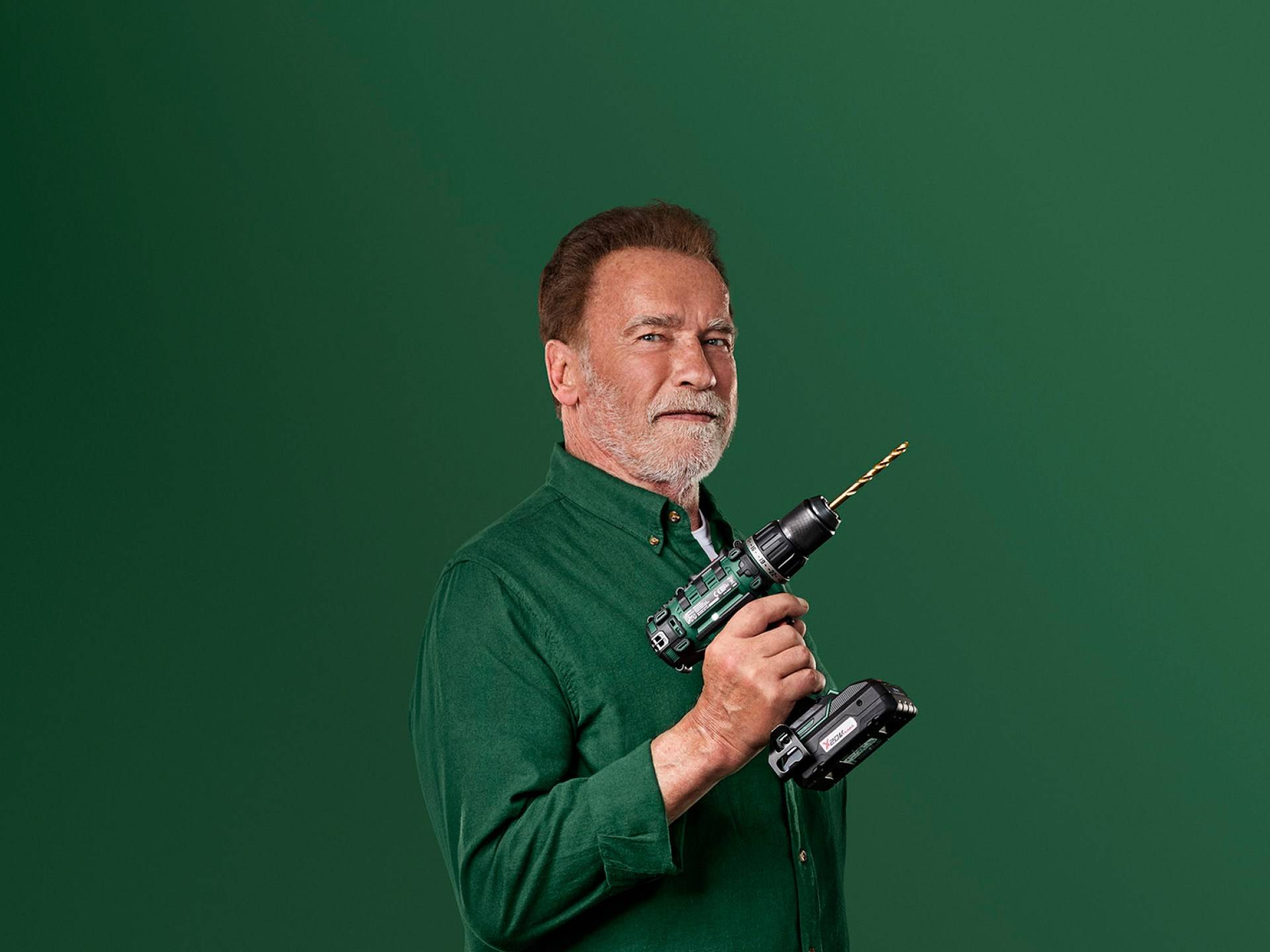 Arnold Schwarzenegger houdt een groene elektrische boormachine vast tegen een groene achtergrond.