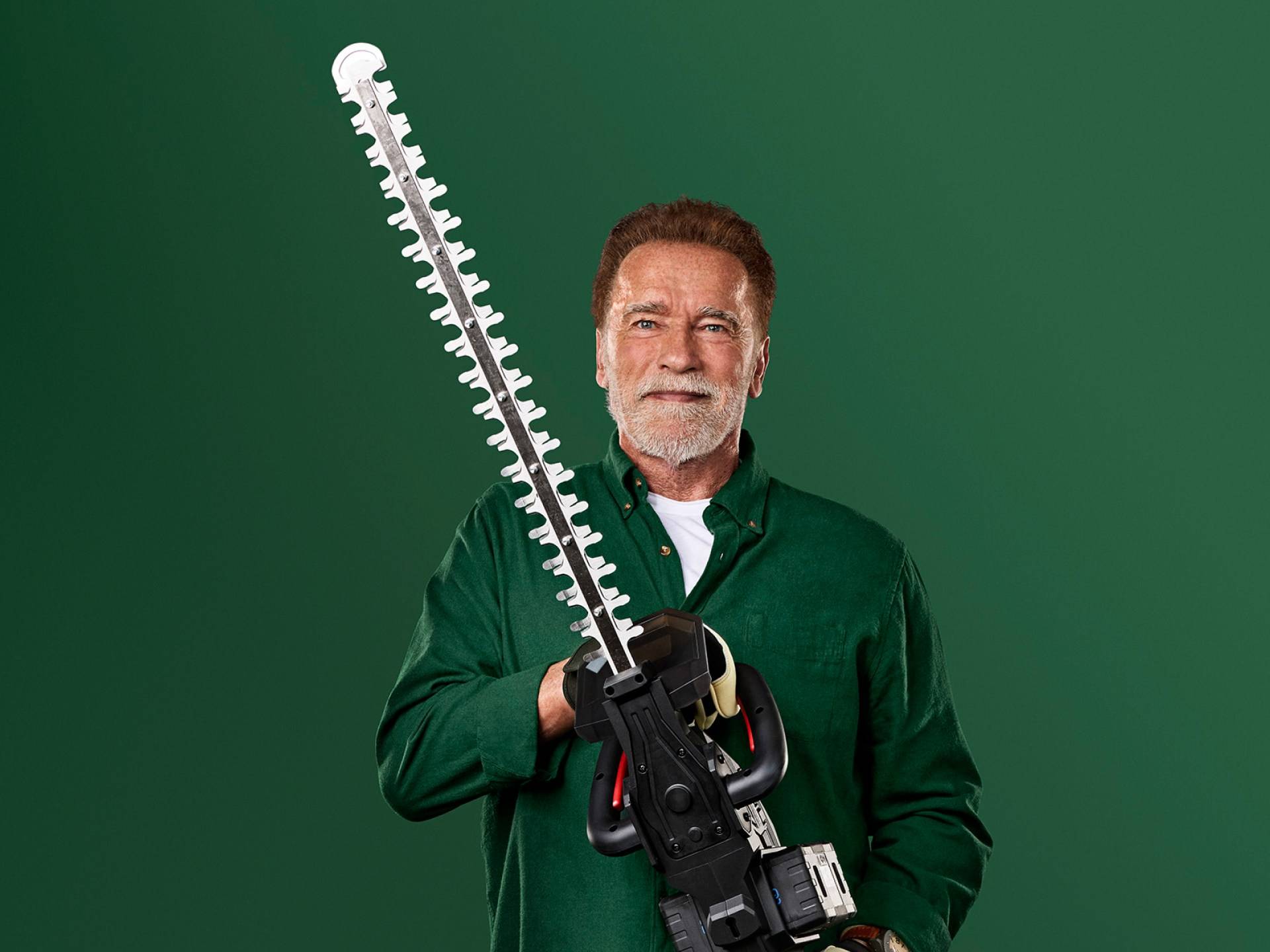 Arnold Schwarzenegger houdt een grote heggenschaar vast tegen een groene achtergrond.