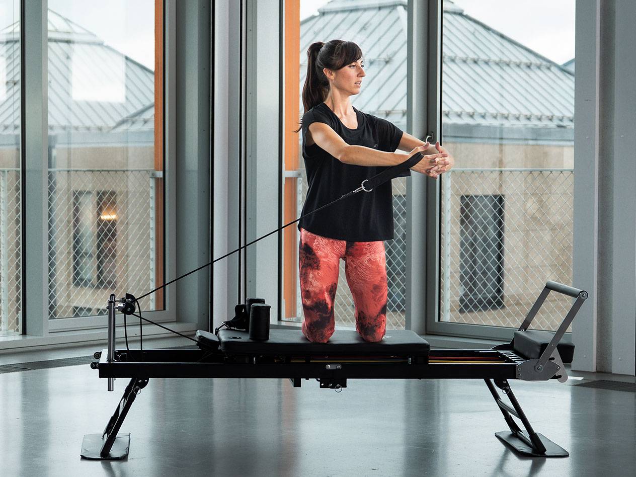 Vrouw doet Pilates op een zwarte reformer machine in een lichte kamer.