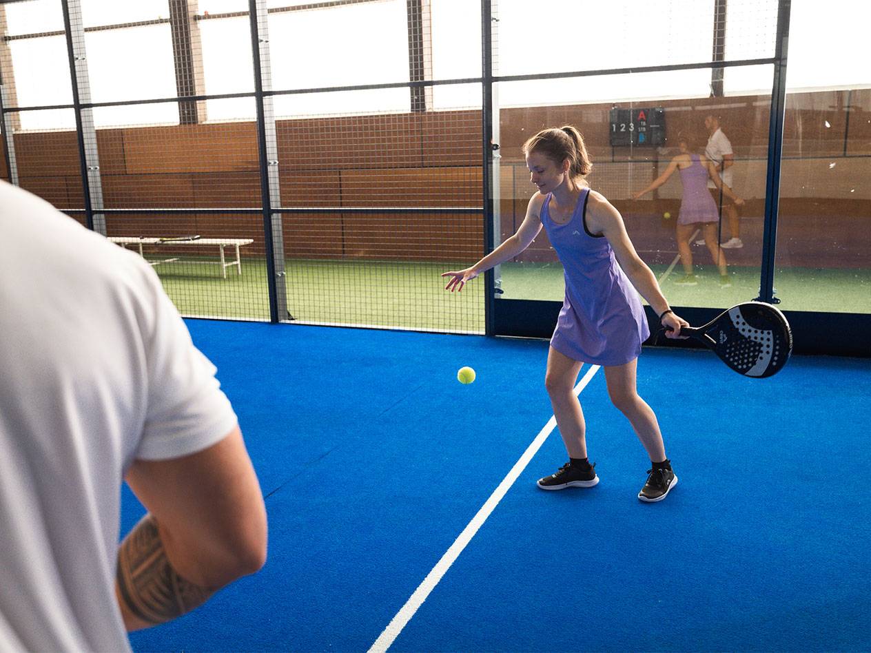 Vrouw speelt padel, houdt racket vast en kijkt naar de bal op een blauw veld.