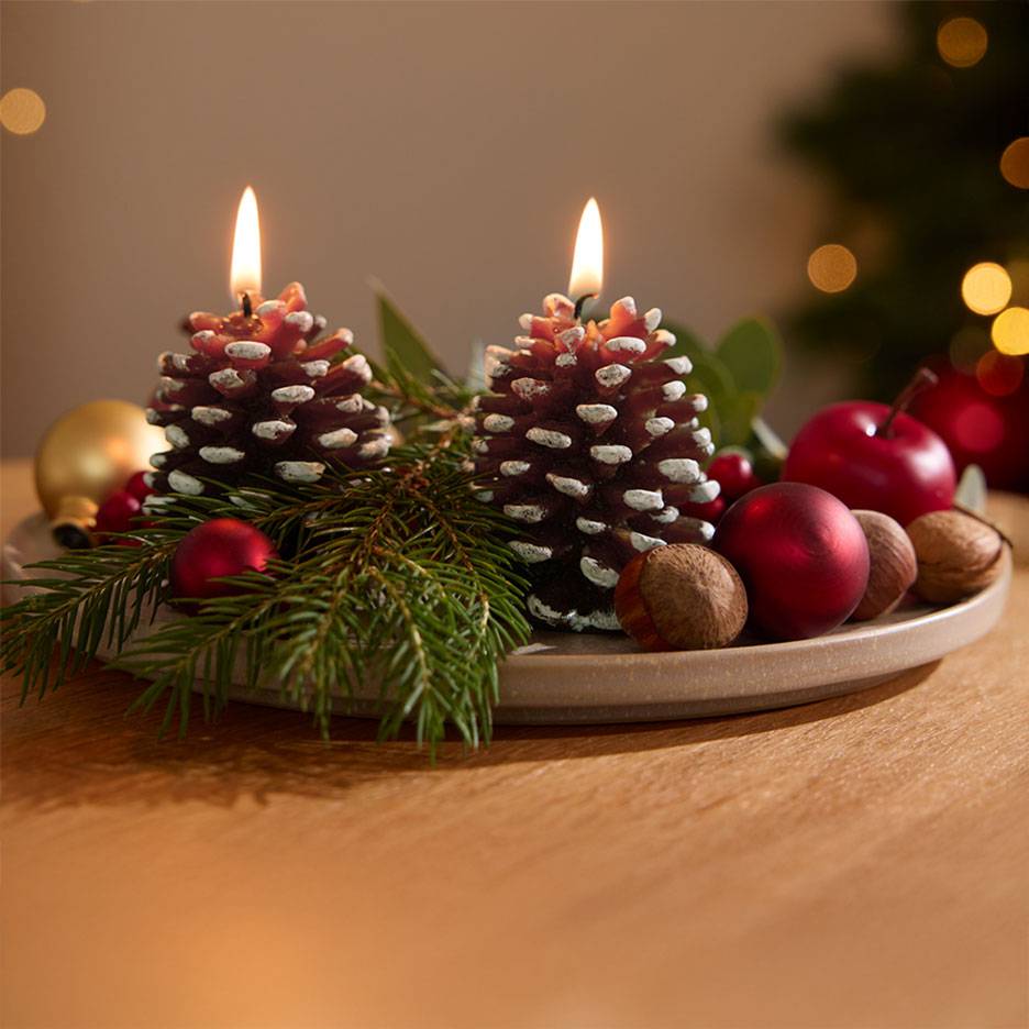 Adventsdecoratie met twee brandende dennenappelkaarsen, dennentakken en kerstversiering op een bord.