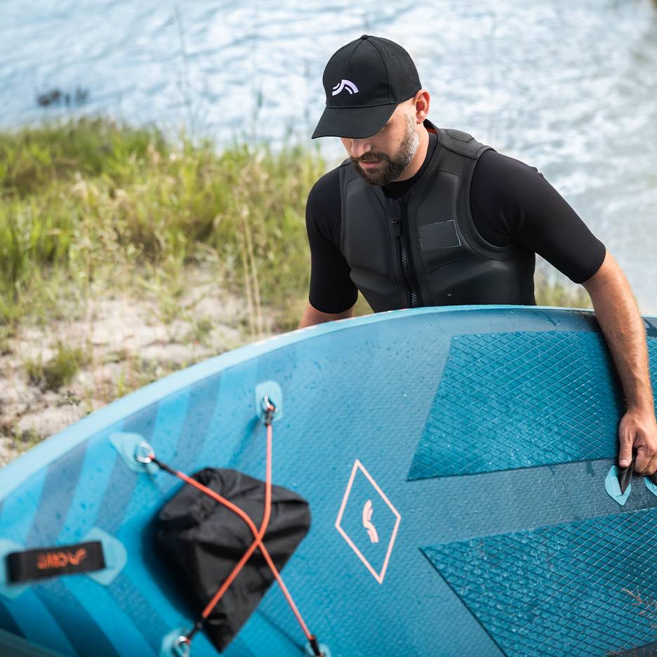 Man met zwemvest en pet houdt blauw paddleboard vast bij het water.