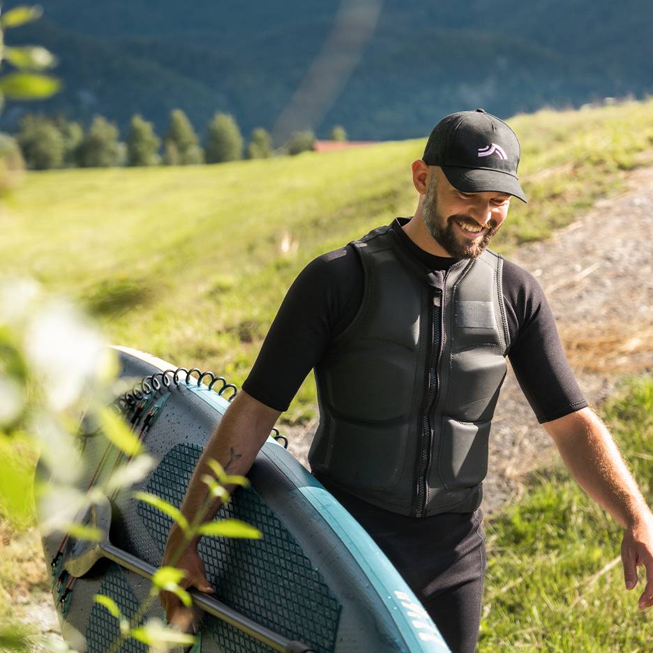 Man in zwart wetsuit en zwemvest, draagt een SUP-board met peddel.