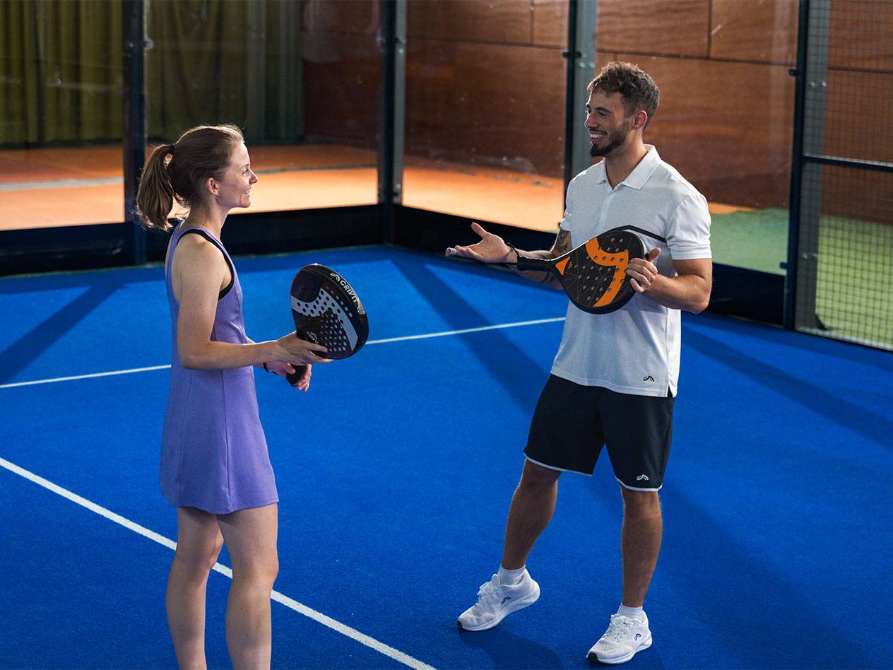Twee padelspelers met rackets op een blauw veld, lachend naar elkaar.