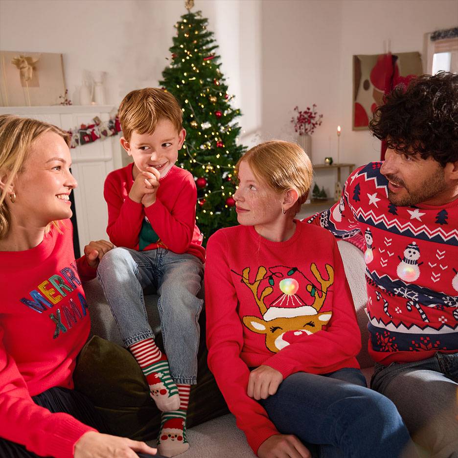 Familie in rode kersttruien met rendieren en sneeuwpoppen, zittend op een bank voor een kerstboom.