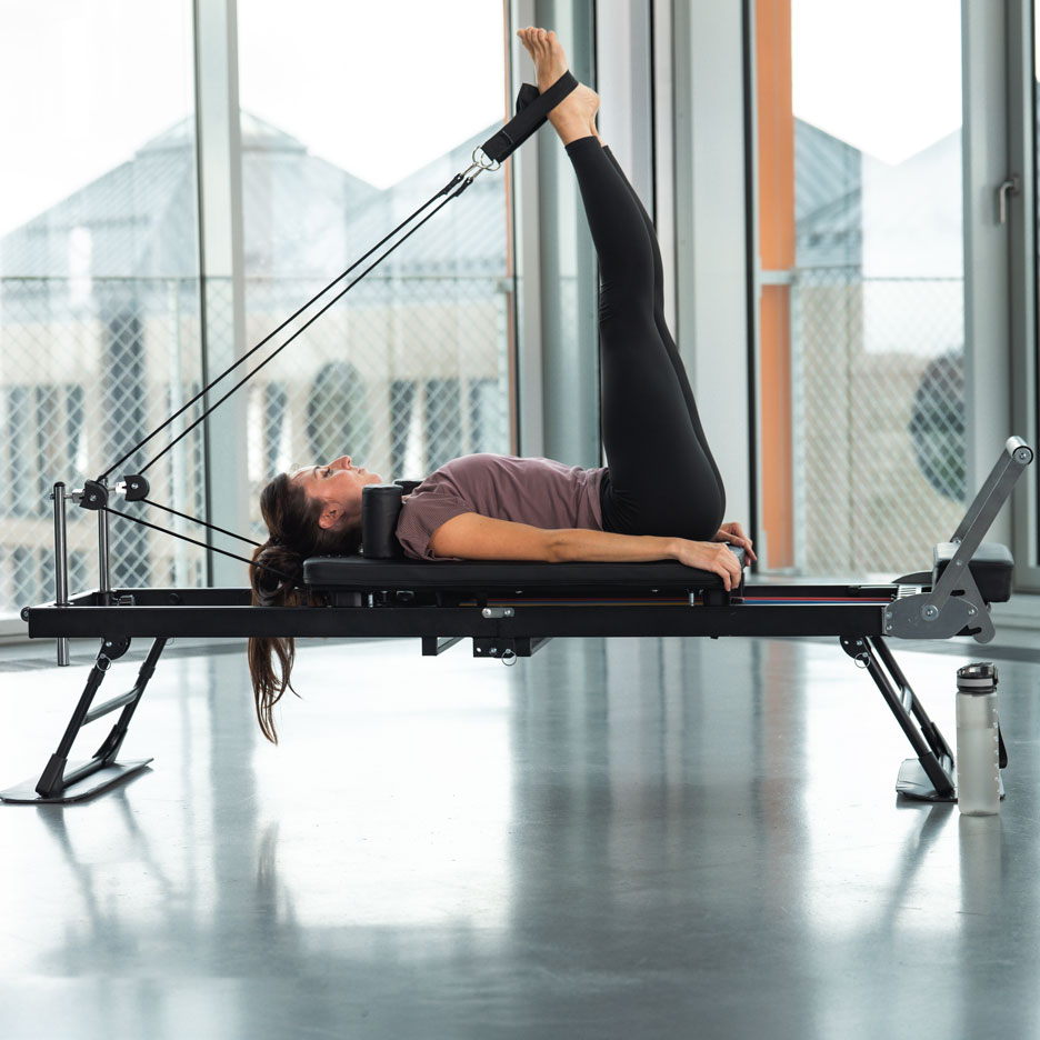 Vrouw doet Pilates op een reformer met benen omhoog, in een lichte kamer.