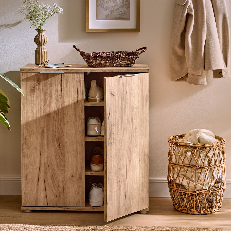 Houten schoenenkast met open deur, schoenen, vaas met bloemen en manden in een hal.
