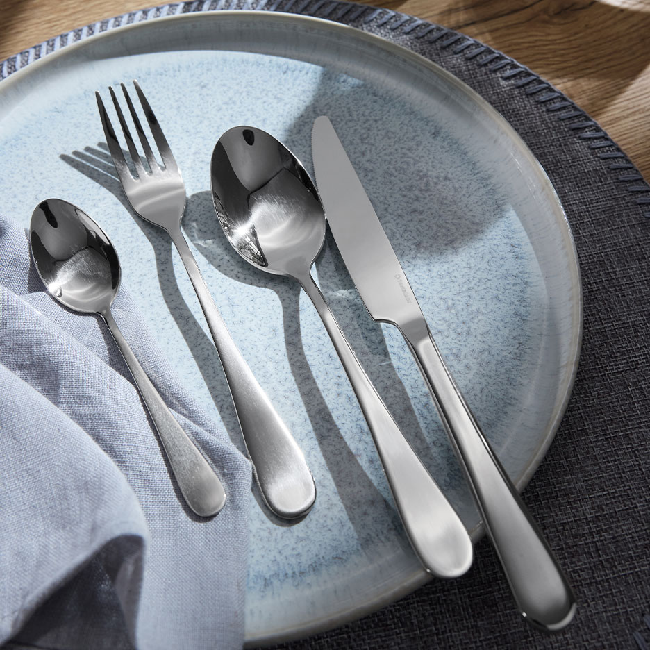 Ensemble de couverts brillants : fourchette, cuillère, petite cuillère et couteau sur une assiette bleue.