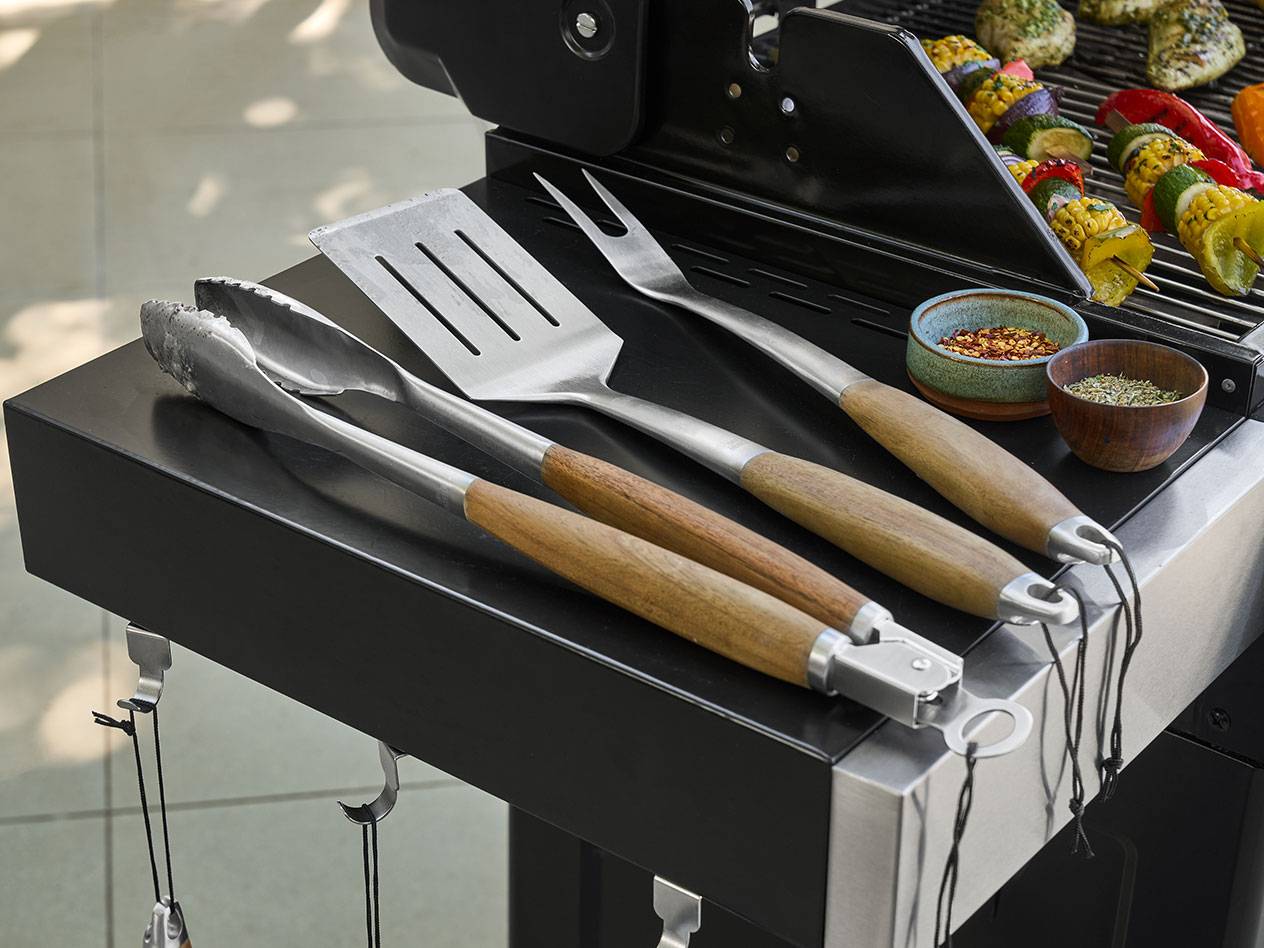 Barbecuegereedschap met houten handvatten en kruiden naast gegrilde groentespiesjes.