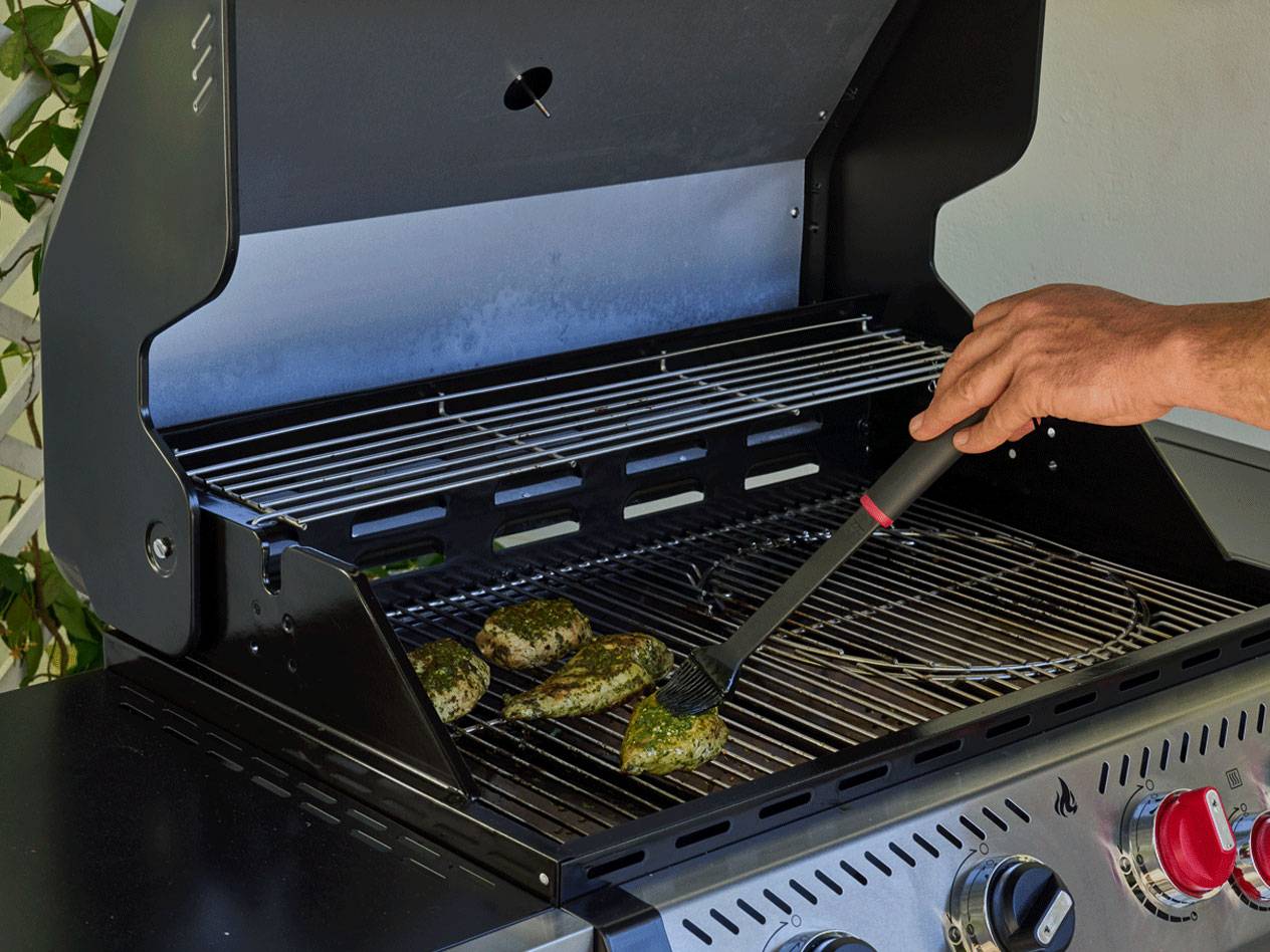 Een persoon die gekruide kipstukken op een gasbarbecue bestrijkt met een kwast.