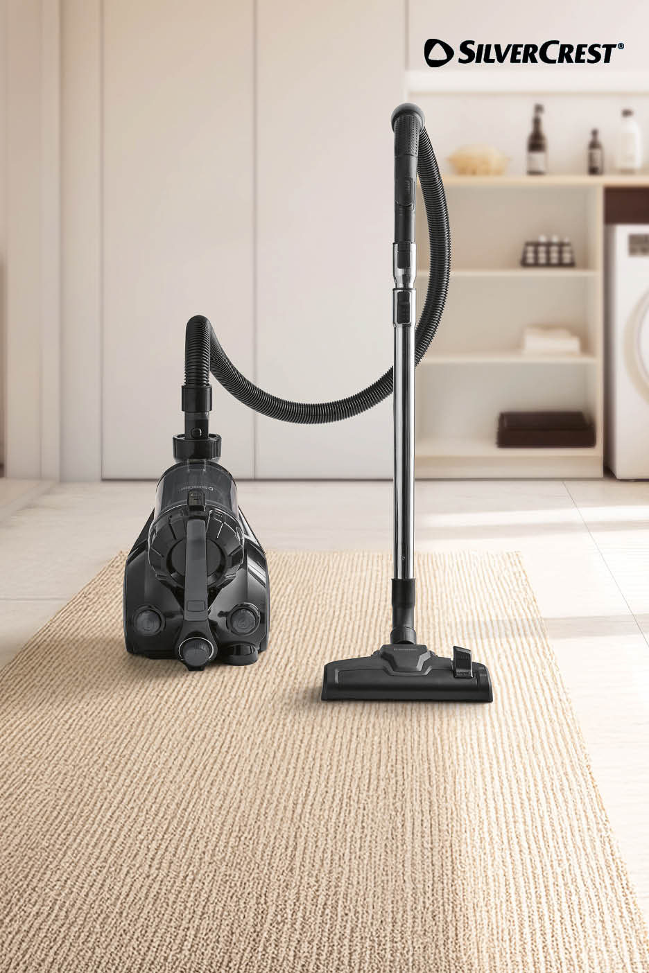 Een zwarte zakloze stofzuiger op een beige tapijt in een lichte kamer.