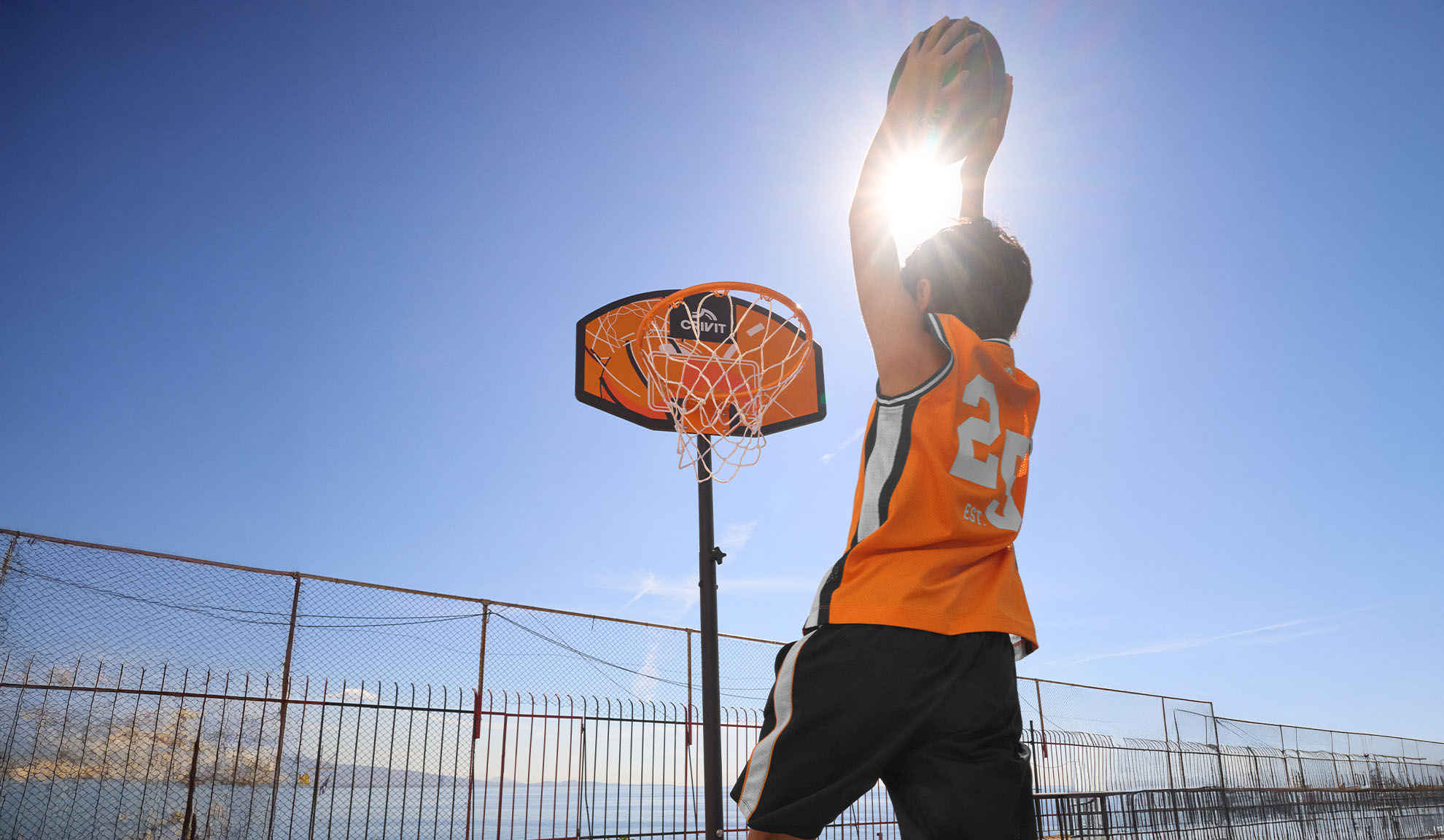 Enfant en maillot de basket tirant au panier en extérieur sous un soleil éclatant