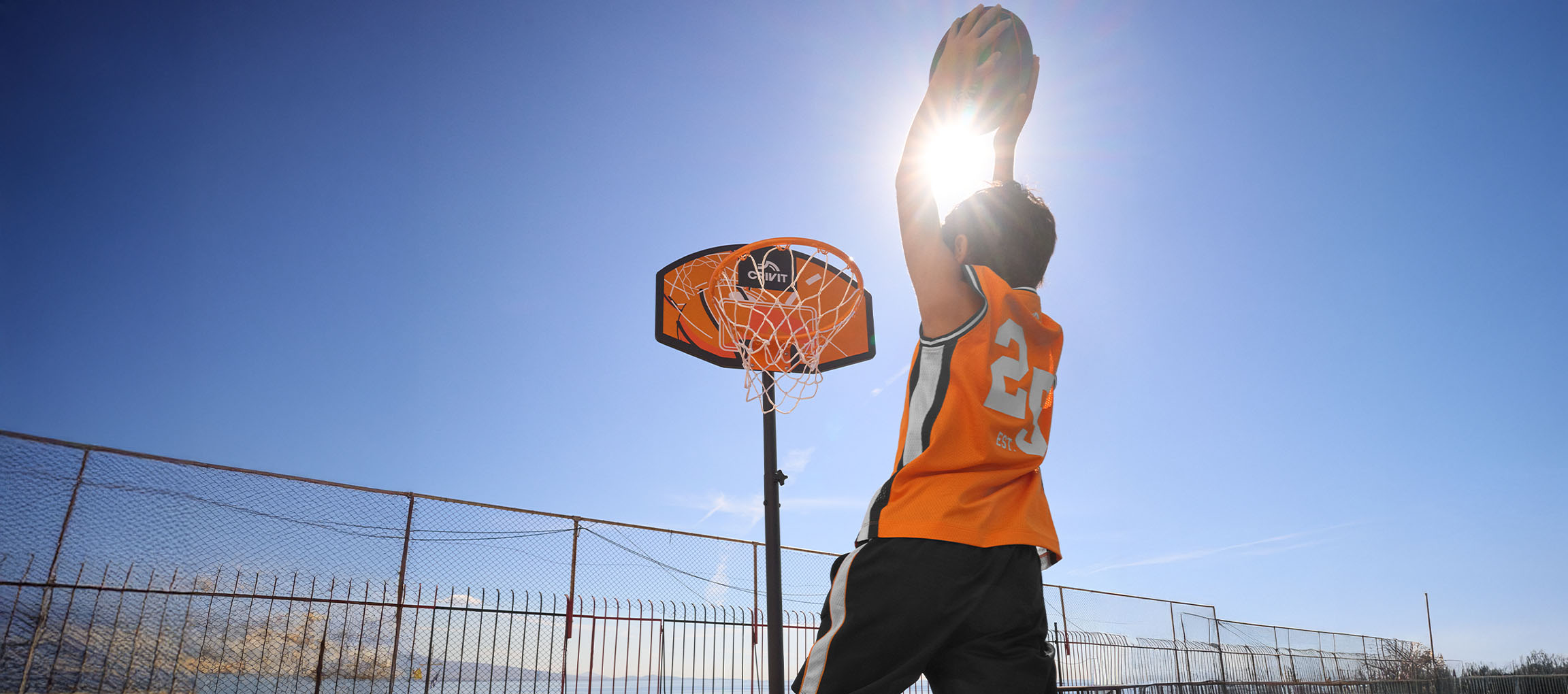 Jongen in oranje basketbalshirt gooit bal in de basket op een zonnige dag.