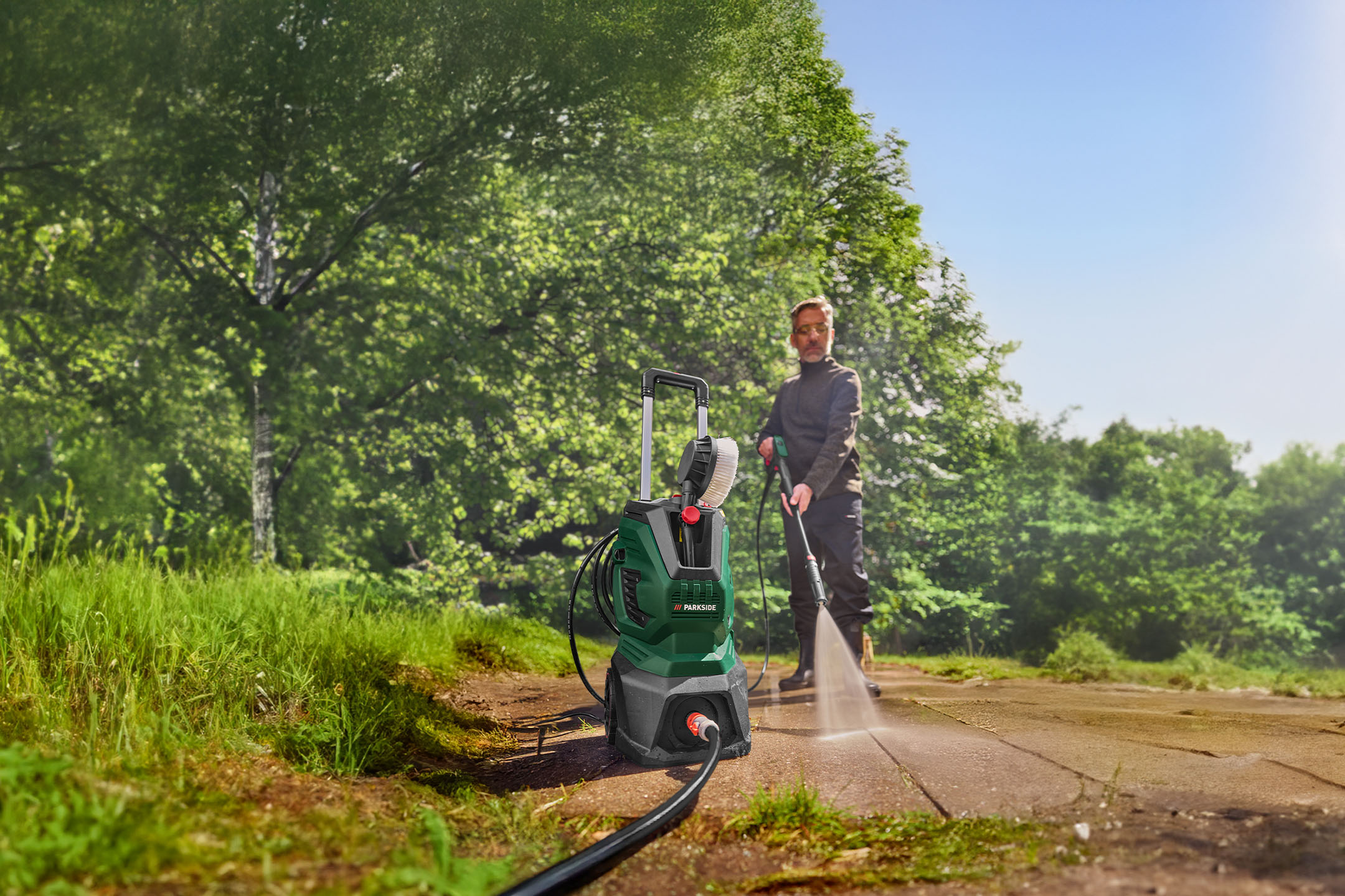 Homme nettoyant un chemin avec un nettoyeur haute pression et une brosse rotative dans un jardin.