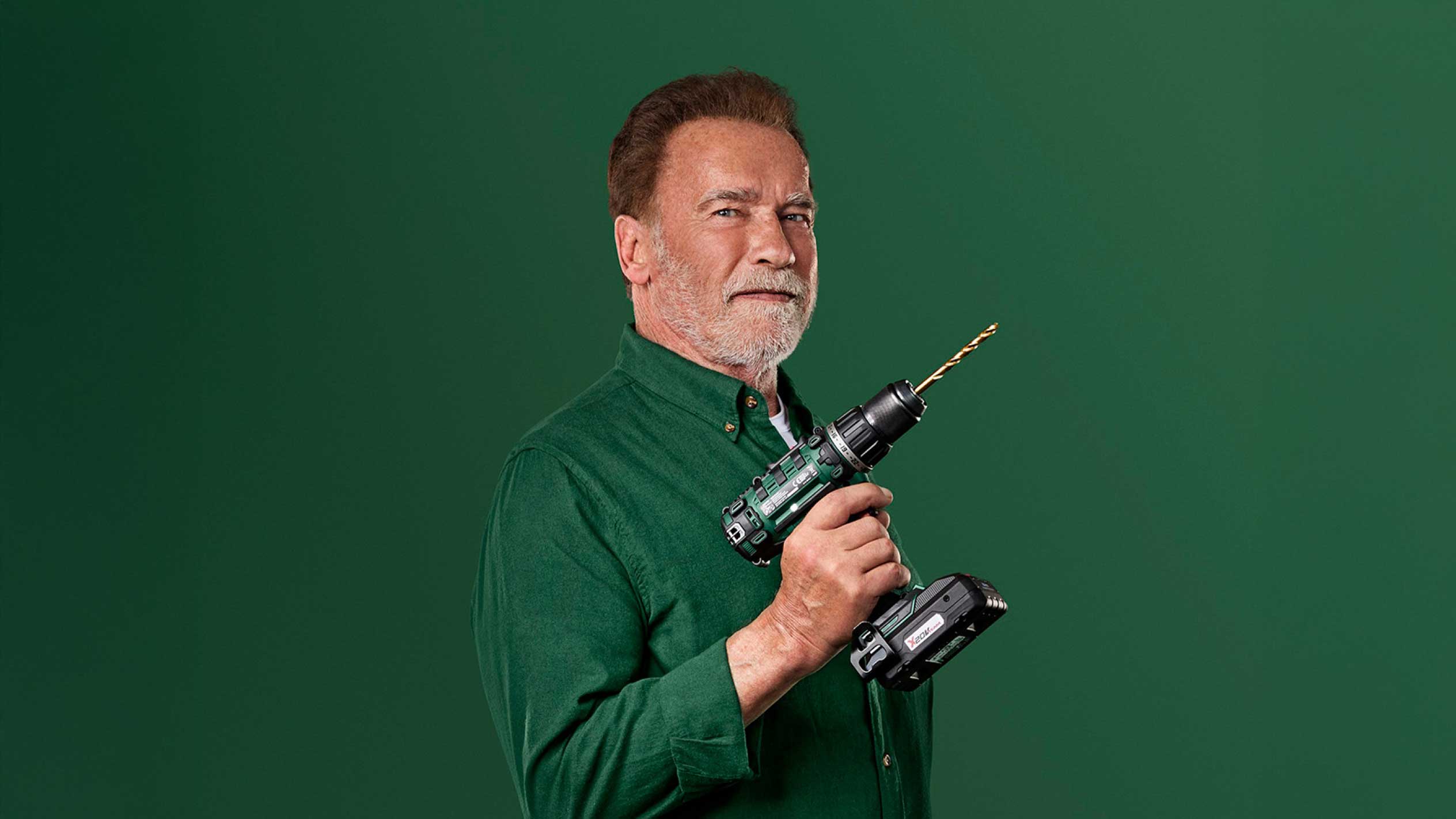 Arnold Schwarzenegger houdt een groene accuboormachine met een gouden boor vast.