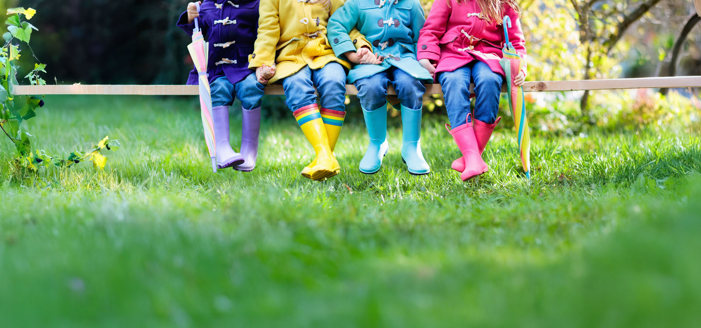 Kinderen in kleurrijke regenjassen en laarzen zitten op een bankje in het gras.
