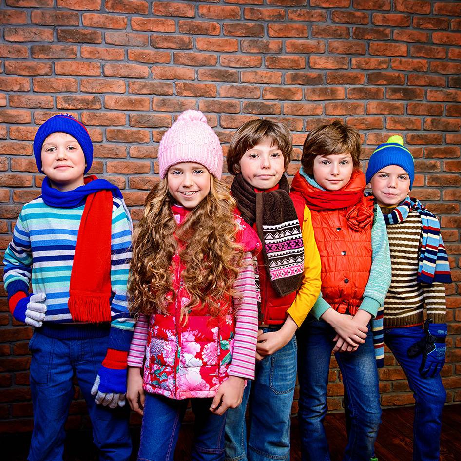 Vijf kinderen in kleurrijke wintermutsen, sjaals en vesten voor een bakstenen muur.