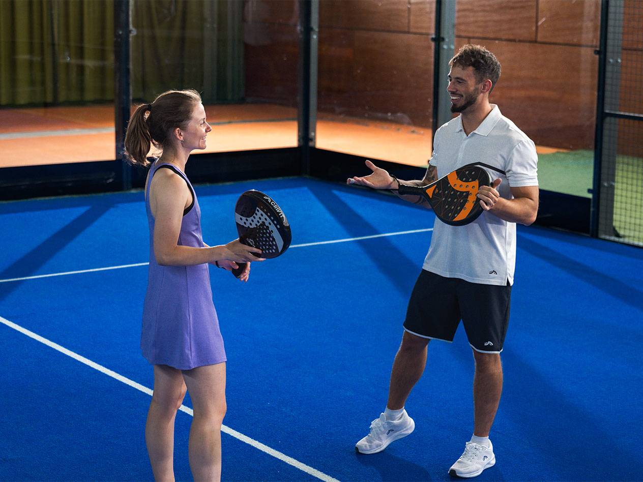 Twee mensen op een padelbaan, met padelrackets in de hand en in gesprek.