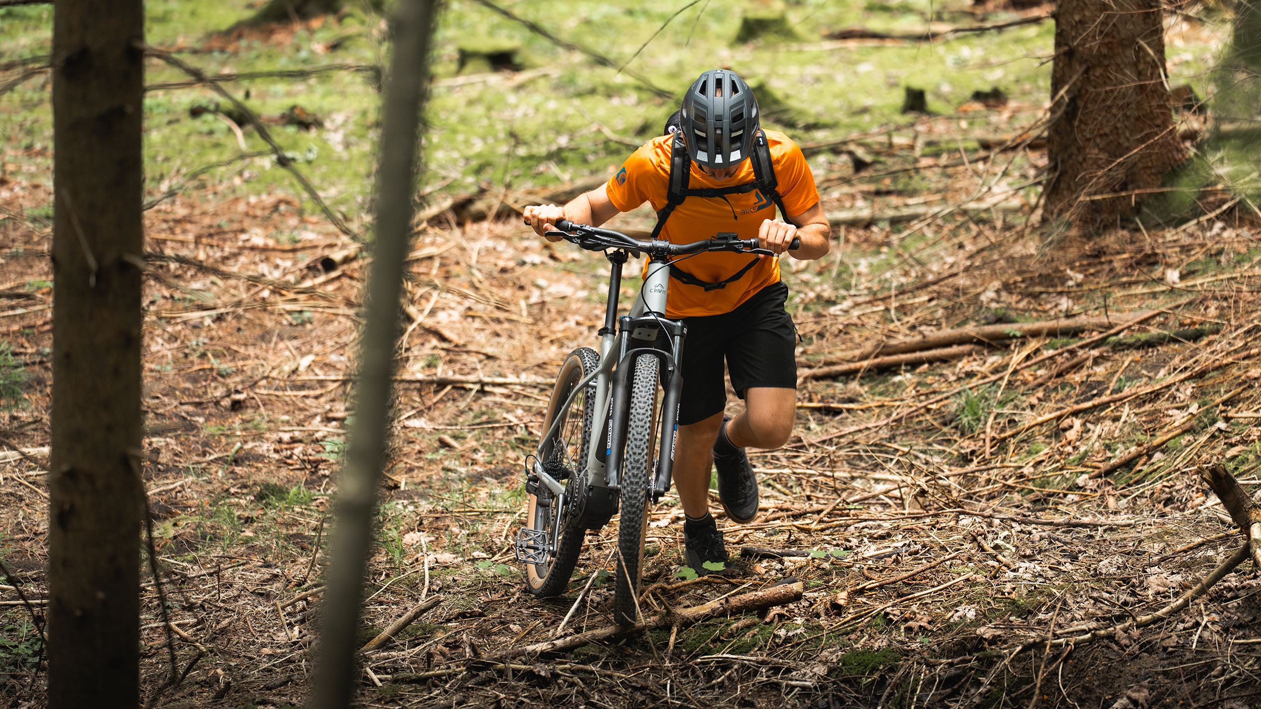 Een fietser in een oranje shirt duwt een elektrische mountainbike door een bos.