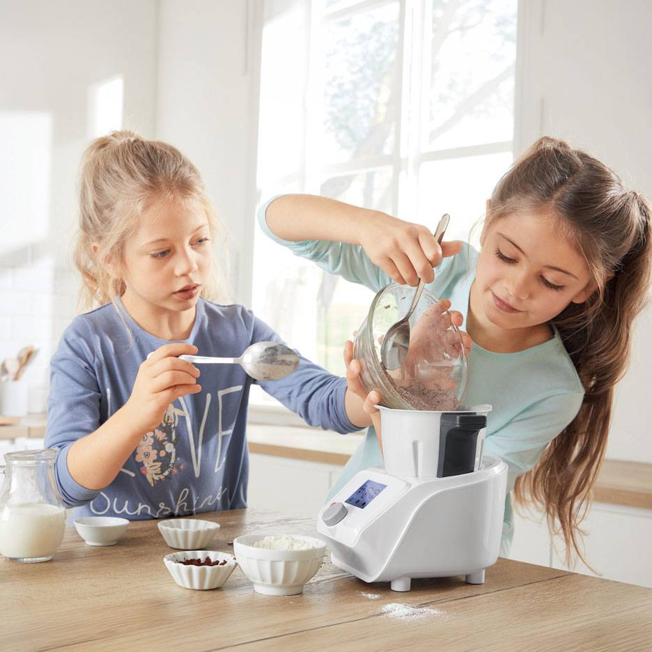 Twee meisjes bereiden deeg in een keukenmachine, met kommen ingrediënten op tafel.