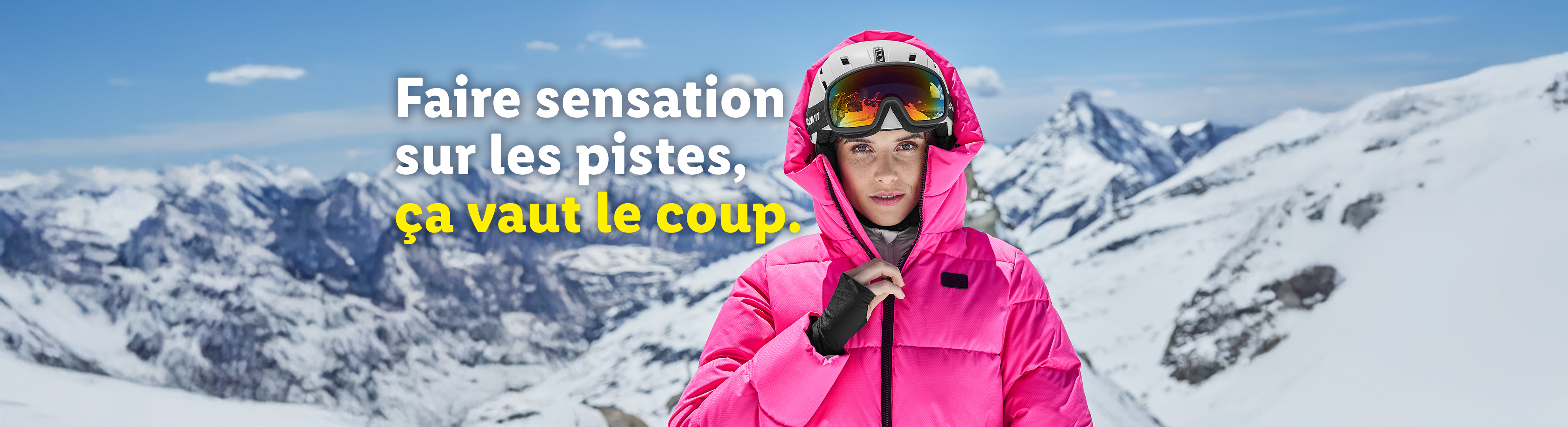 Femme en veste de ski rose et casque avec masque de ski, montagnes enneigées en arrière-plan.