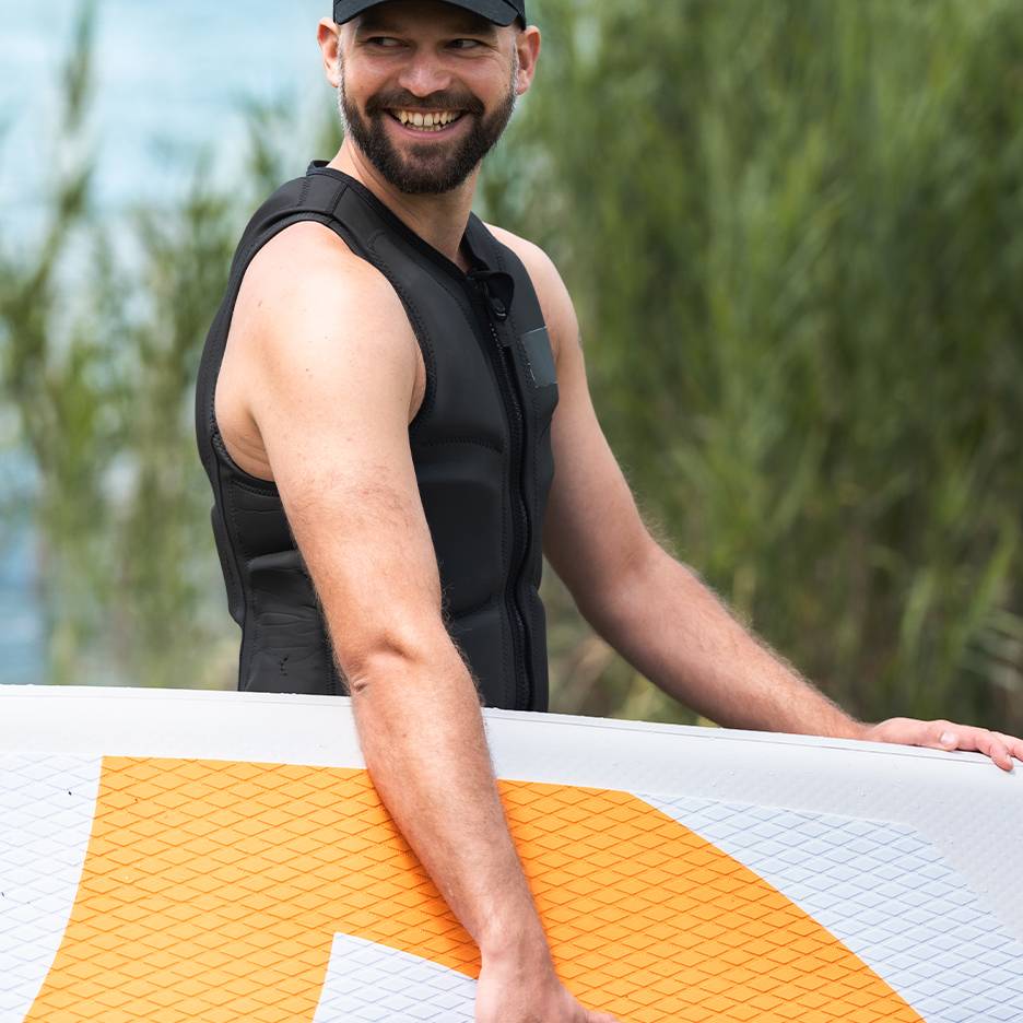 Lachende man in zwart zwemvest houdt een paddleboard vast.