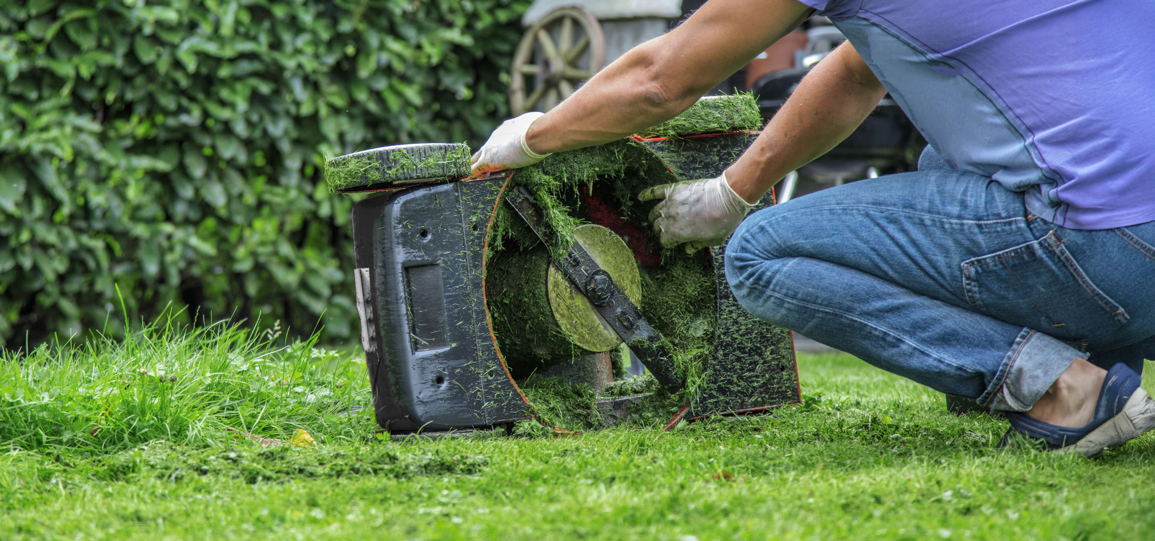 Personne nettoyant une tondeuse à gazon couverte d'herbe fraîchement coupée dans un jardin.
