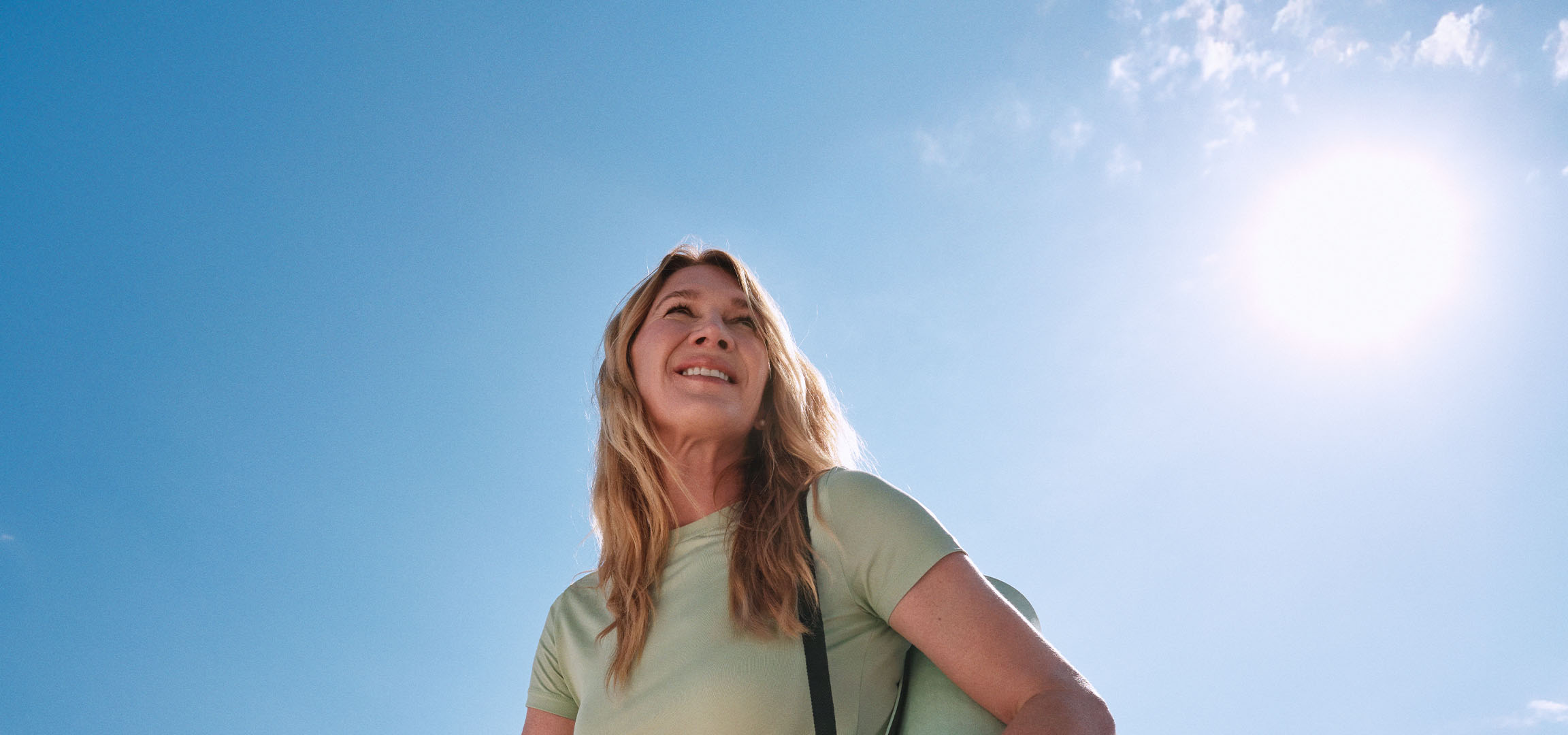 Lachende blonde vrouw kijkt naar de zon onder een open hemel.