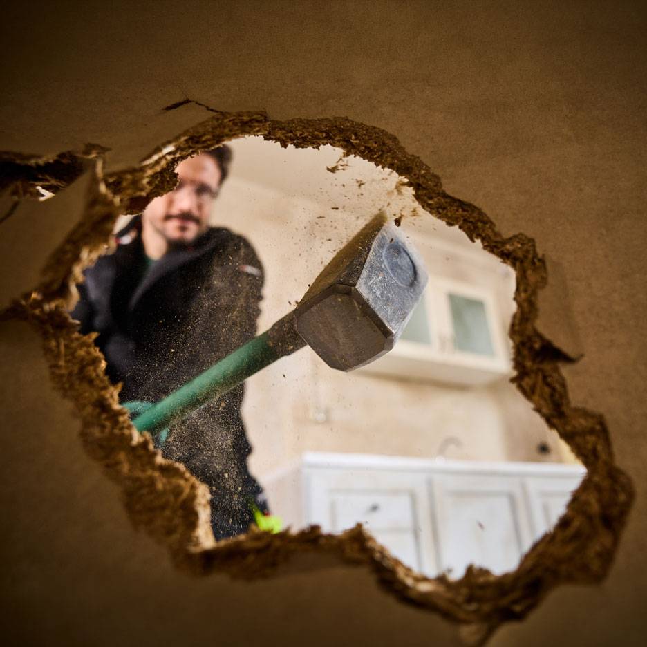 Homme avec masse démolissant un mur, vu à travers un trou, avec des armoires de cuisine en arrière-plan.