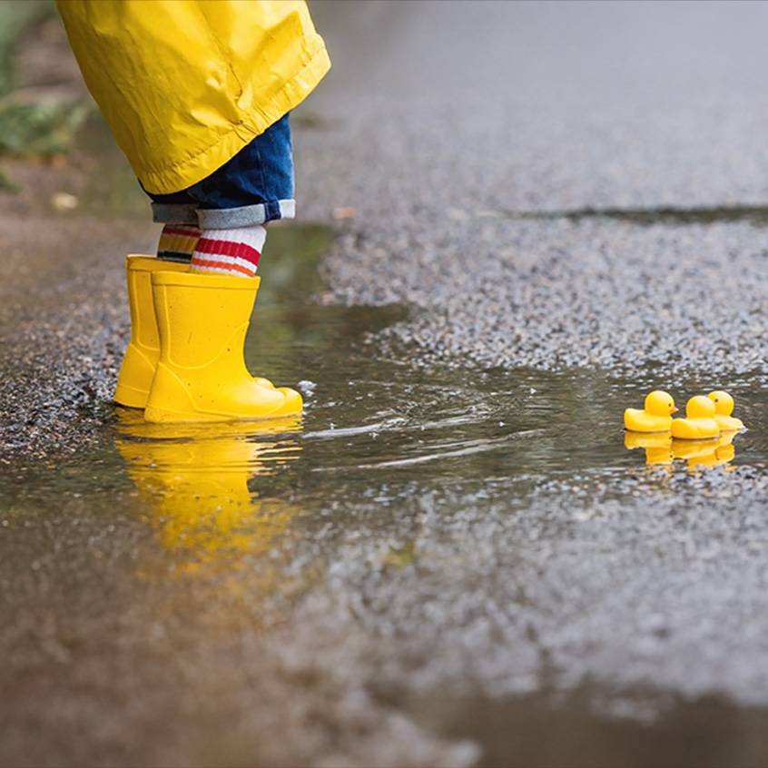 Kind in gele regenlaarzen en jas in een plas met badeendjes.