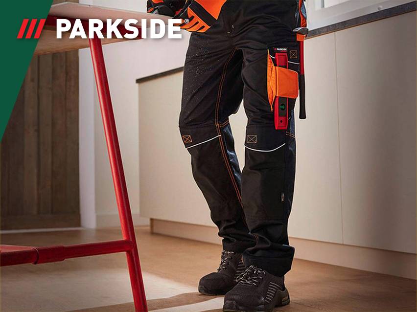 Werkbroek met oranje accenten en gereedschap, gedragen door een persoon in een interieur.