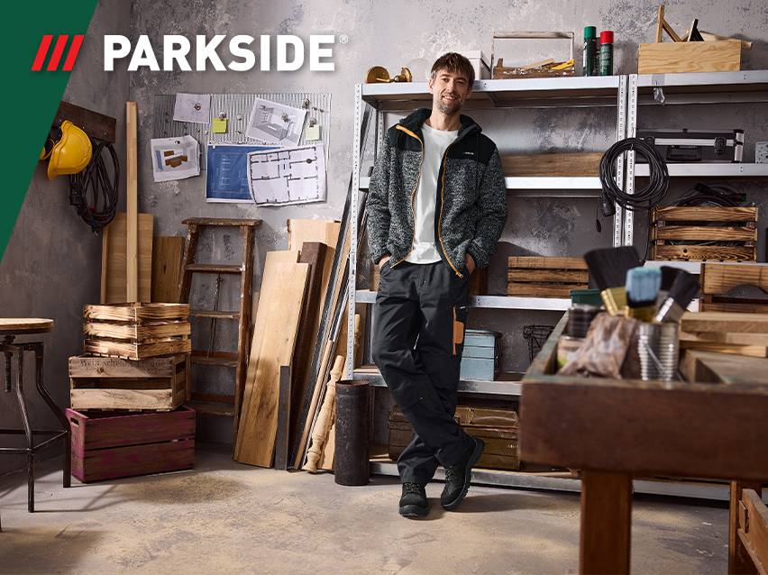 Man in werkplaats met gereedschap en hout, draagt een fleecejack en werkbroek.