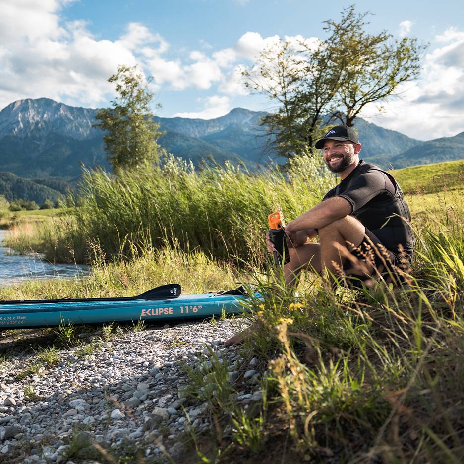 Lachende man met pet en zwemvest, zittend bij een blauw paddleboard aan de rivier.
