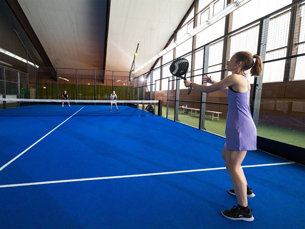 Vrouw in paarse jurk speelt padel op een blauwe baan, met een racket.