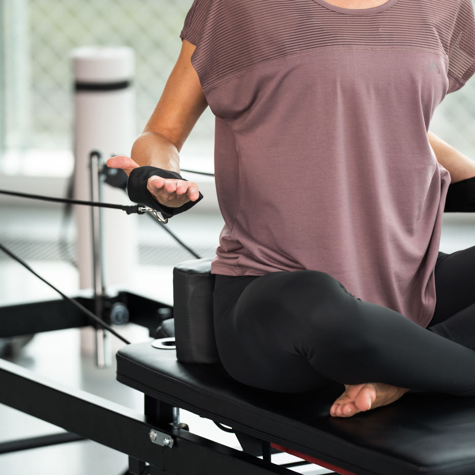 Vrouw doet Pilates op een reformer, draagt zwarte legging en een paarse top.
