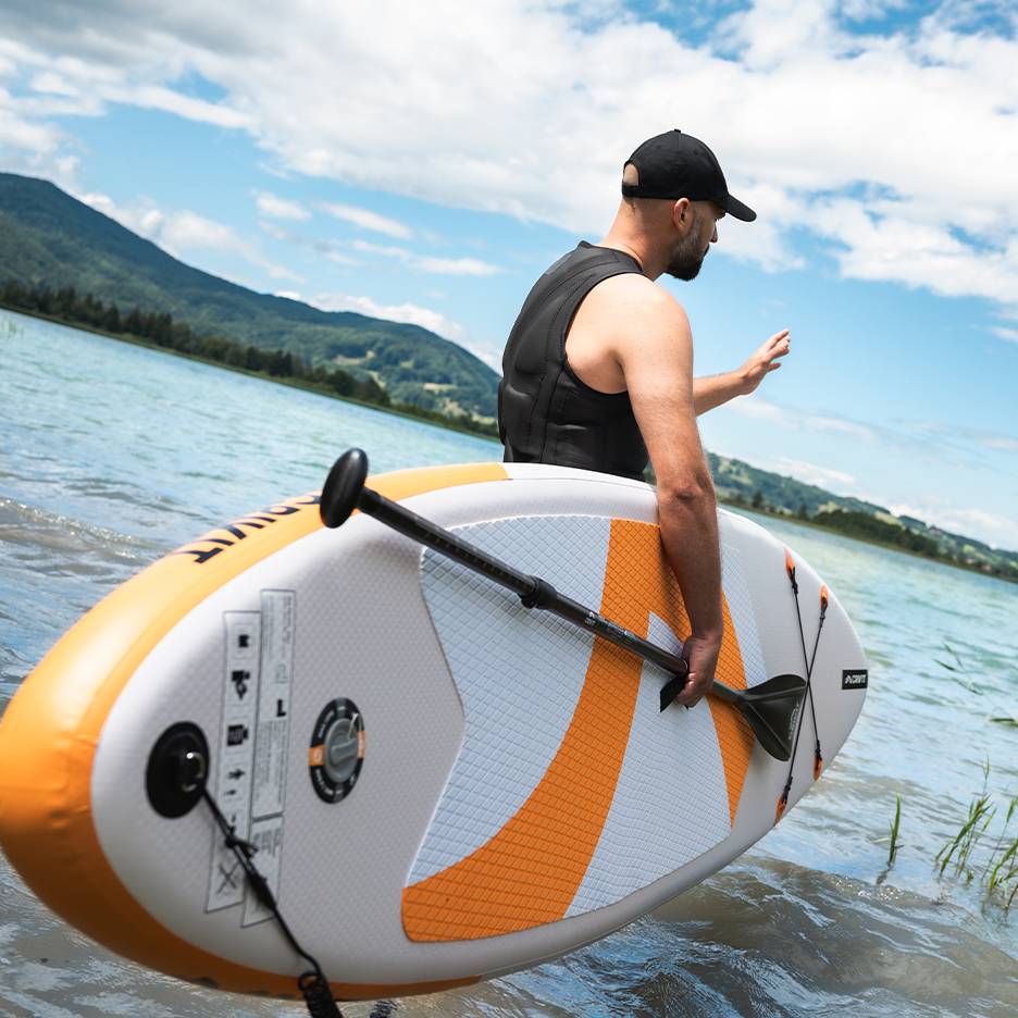 Man met zwemvest en pet houdt opblaasbaar paddleboard in het meer.