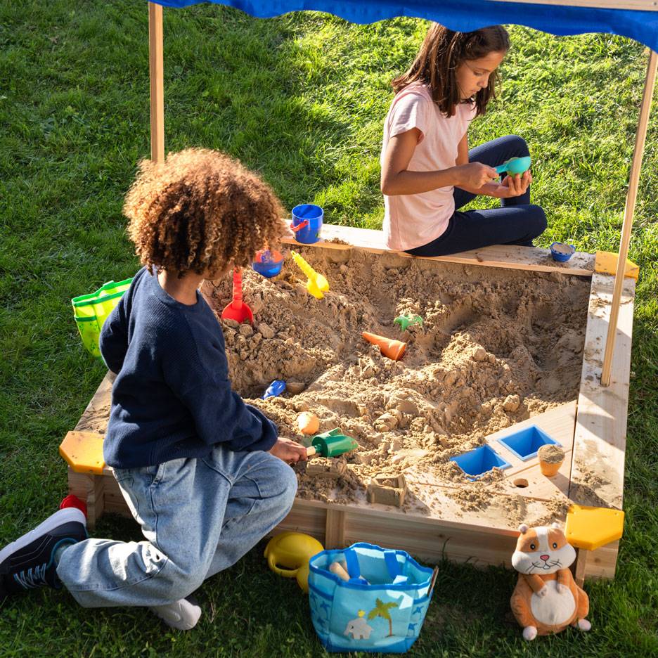 Twee kinderen spelen in een zandbak met luifel, omringd door zandspeelgoed.