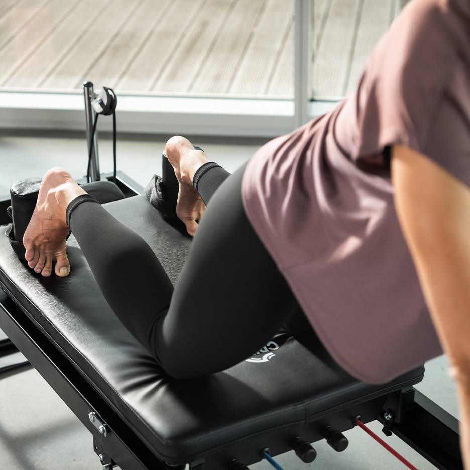 Vrouw traint op een zwarte Pilates-machine met voeten in riemen.