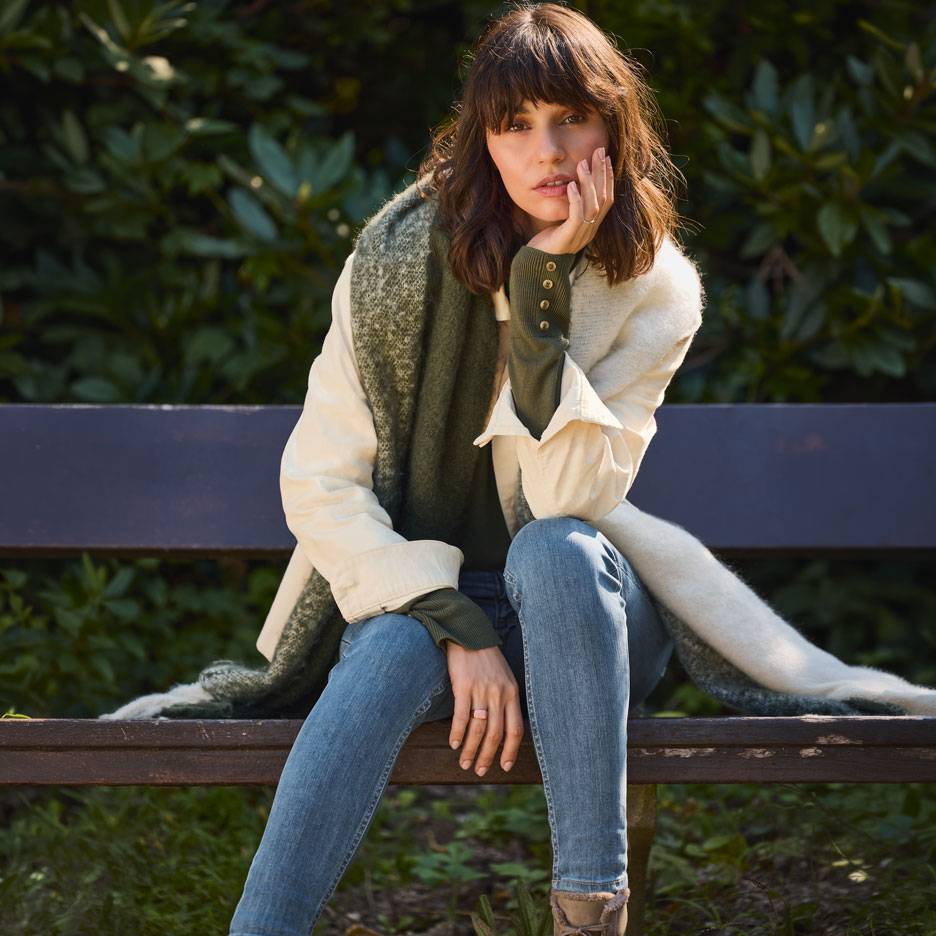 Vrouw in jeans, witte jas en groene sjaal zittend op een parkbankje.