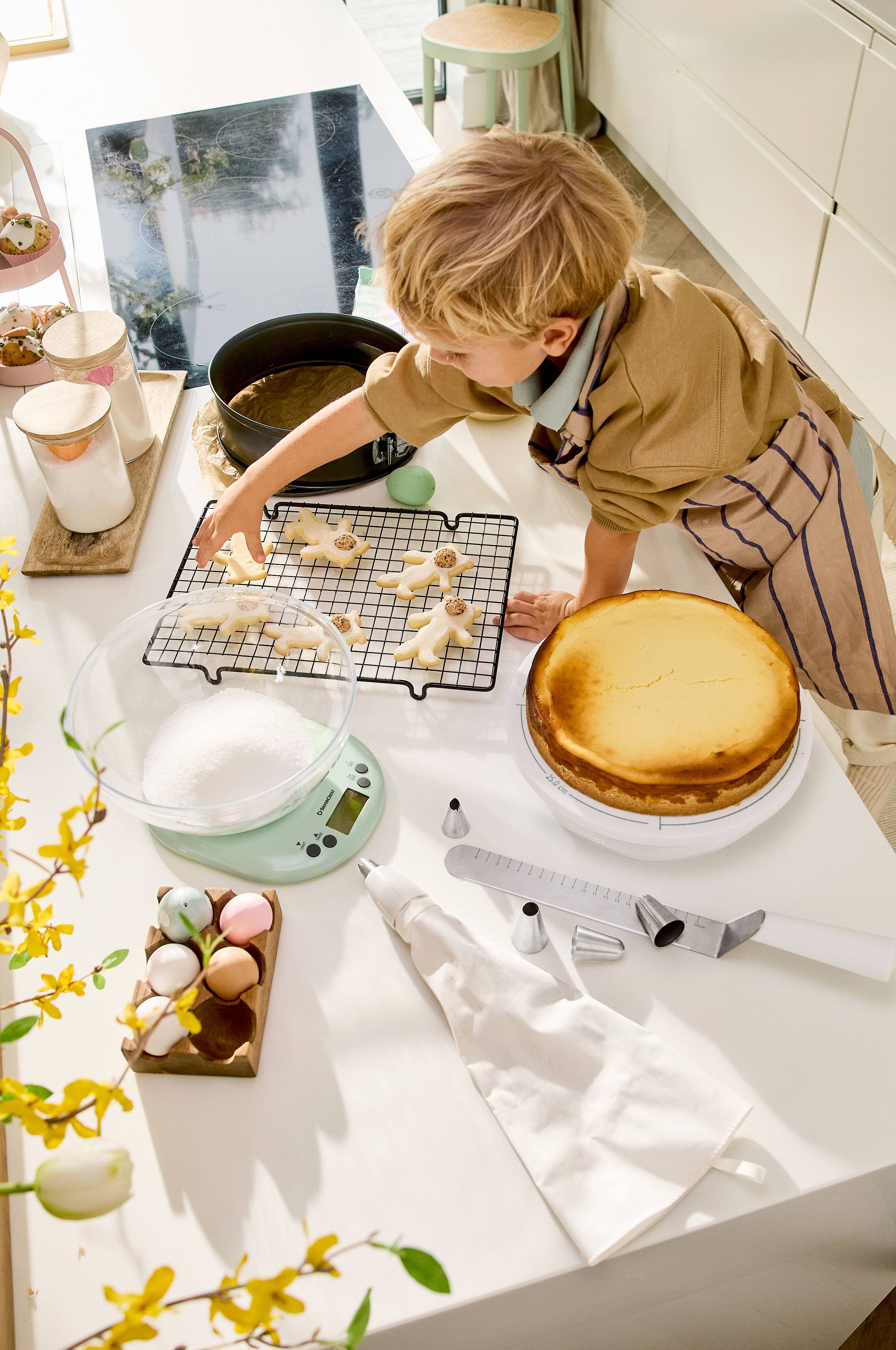 Een kind versiert paaskoekjes, met een cheesecake en bakgerei op tafel.