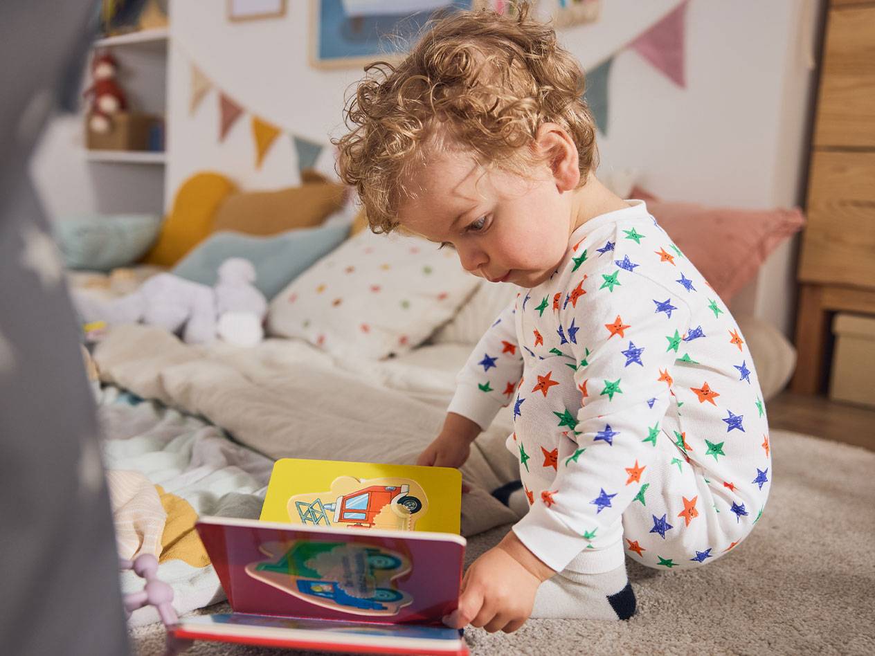 Een peuter in sterrenpyjama, zittend op het vloerkleed, leest een boek.