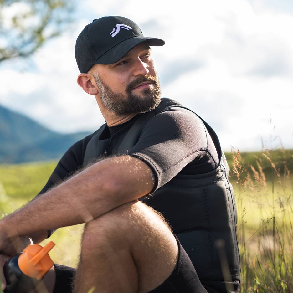 Man met zwarte pet en neopreen vest zit in het gras, kijkt opzij.